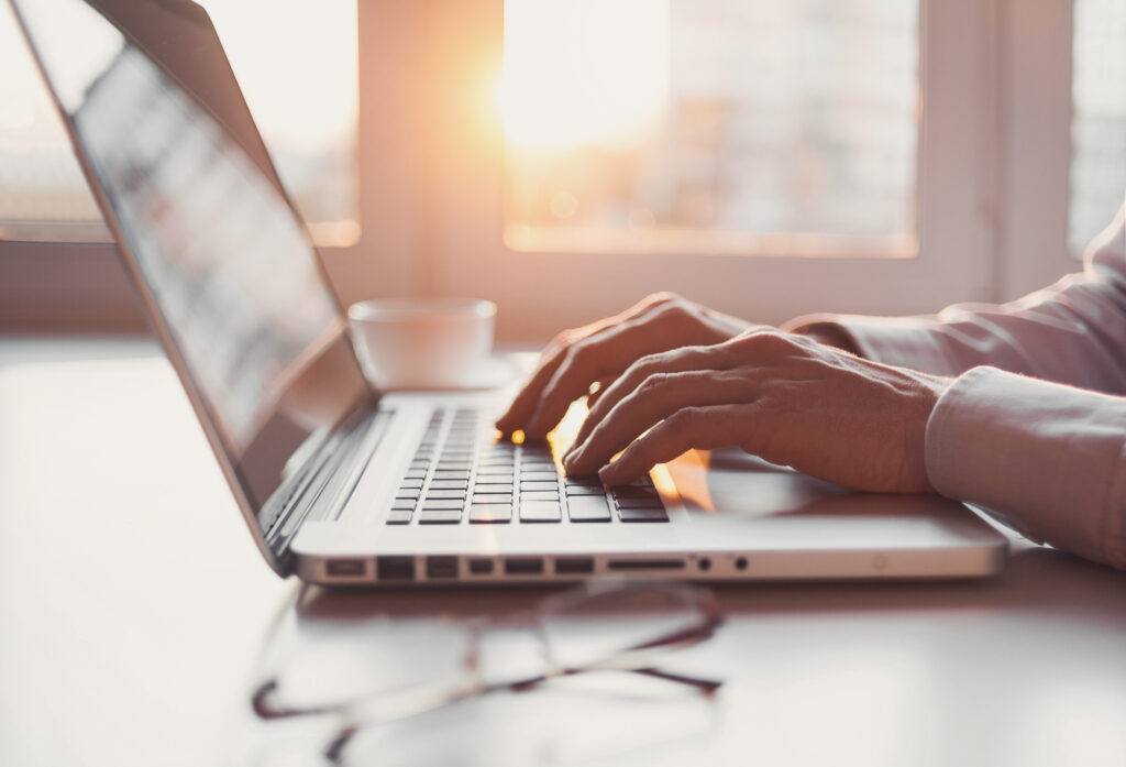 Hands typing on a laptop keyboard at a sunlit desk with a coffee cup and glasses nearby.